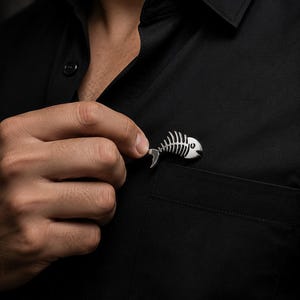 May include: A polished silver-toned metal lapel pin shaped like a fish skeleton. The pin features a detailed head, a curved spine, and rib-like structures, attached to a solid black button-down shirt with a visible chest pocket and collar.