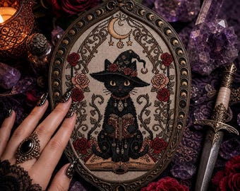 May include: An oval-framed cross-stitch embroidery featuring a black cat wearing a witch hat adorned with roses, reading a book. The cat is surrounded by floral vines and a crescent moon. The frame has a metallic finish with moon phase details.