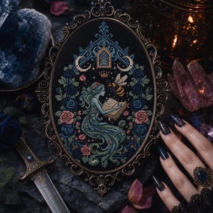 May include: An oval-framed cross-stitch embroidery on black fabric featuring a central figure with long flowing hair reading a book, surrounded by a bee, a crown, and floral motifs in shades of blue, green, and pink, encased in an ornate metal frame.