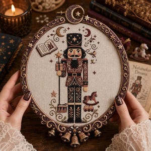 May include: A circular embroidery hoop frame containing a cross-stitch design of a nutcracker soldier in brown and black tones. The design features a crescent moon, stars, an open book, a rocking horse, and a drum. The frame is decorated with small bells.