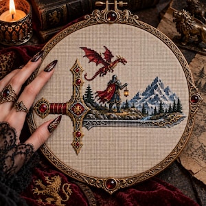 May include: A circular embroidery hoop containing a cross-stitch design of a sword with a red dragon flying above it. A figure in a red cloak holds a lantern near a mountain range and pine trees. The hoop has an ornate, metallic gold frame.