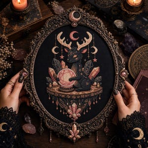 May include: An oval-framed cross-stitch embroidery on black fabric featuring a dark deer with antlers holding a pink crystal ball. The design includes surrounding crystals, stars, and crescent moon motifs, all encased in an ornate, dark metallic frame.