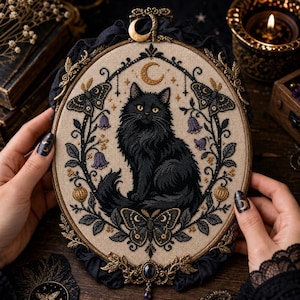 May include: An oval embroidery hoop containing a cross-stitch design of a fluffy black cat centred within a wreath of purple bellflowers, moths, and a golden crescent moon. The hoop features an ornate gold metal frame with a crescent moon topper.
