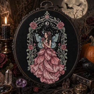 Peut inclure: Une broderie au point de croix ovale représentant une fée centrale avec des ailes bleues et une robe rose à volants, encadrée par une arche florale complexe avec des roses et un croissant de lune sur un fond en tissu noir uni.