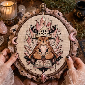 May include: A circular cross-stitch embroidery featuring a brown creature wearing a witch hat with moon phases, holding a crystal ball above an open book. The design is framed by pink crystals, black stars, and crescent moons on beige fabric.
