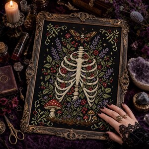 May include: A framed cross-stitch embroidery on black fabric featuring a human rib cage surrounded by colourful wildflowers, red mushrooms, and a moth with a skull pattern on its back, all enclosed in an ornate, antique-style gold metal frame.