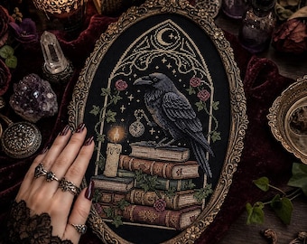 May include: An oval-framed cross-stitch embroidery featuring a black raven perched on a stack of books. The design includes a lit candle, a crescent moon, stars, and small pink roses, all set against a black fabric background within an ornate gold frame.