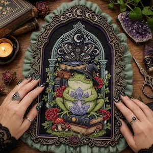 May include: A cross-stitch embroidery piece featuring a green frog wearing a witch hat and holding a purple crystal, surrounded by books, red roses, mushrooms, and a moth, all framed in an ornate, dark metal arch with green fabric trim.