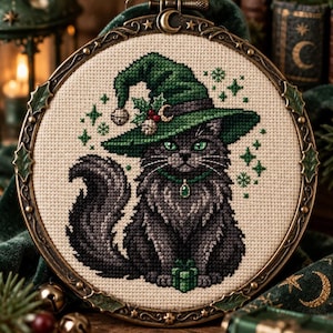May include: A cross-stitch embroidery of a fluffy grey cat wearing a green witch hat adorned with holly berries and a small bell. The cat has green eyes, a green collar with a gem, and holds a small green gift. It is set in a round metal frame.