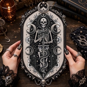 May include: An oval-shaped cross-stitch embroidery piece featuring a central skeleton holding a sword entwined with a snake. The design includes celestial motifs like moons and stars in black, white, and grey threads, framed by a decorative border.