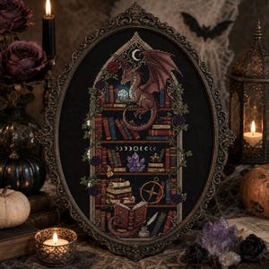 May include: An oval-framed cross-stitch embroidery featuring a dragon perched on a bookshelf filled with books, crystals, and a compass. The design includes a crescent moon, floral vines, and an open book at the base, all rendered in rich colors on black fabric.
