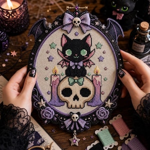 May include: A cross-stitch embroidery piece in an oval black frame with bat wings, featuring a black cat with pink ears and wings sitting on a skull. The design includes two purple candles, a lavender bow with a skull, and small stars.