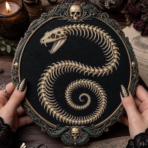 May include: A circular embroidery hoop featuring a cross-stitched beige snake skeleton on a black fabric background. The frame is ornate, dark bronze with teal accents, and decorated with three small skulls at the top, left, and bottom edges.