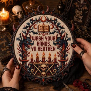 May include: A circular cross-stitch embroidery in a dark ornate frame featuring the text "WASH YOUR HANDS, YA HEATHEN" in red. The design includes two horned creatures, a central open book, five lit candles, and two skulls on a geometric base.