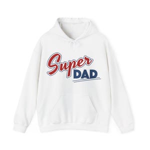 May include: A white long-sleeve hooded sweatshirt featuring a front kangaroo pocket and a drawstring hood. The chest displays the text Super in red script and DAD in bold navy blue block letters, underlined by two thin navy blue horizontal lines.