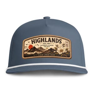 May include: A slate blue baseball cap with a white cord trim along the base of the crown. A rectangular patch features a mountain landscape, a red sun, birds, and a white flower with the text HIGHLANDS EST. 1967 AUTHENTIC FLAVOR FOR BTS CLEAR CREEK GSHR.
