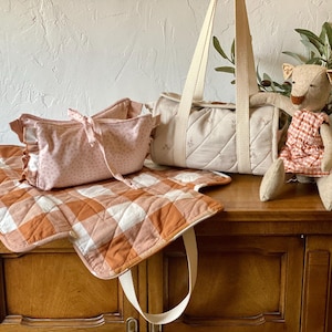 May include: A quilted diaper bag with cream-colored straps, a matching pink patterned pouch with ruffled edges, a folded orange and white gingham changing mat, and a stuffed animal wearing a matching orange and white gingham dress.