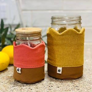 Puede incluir: Dos frascos de vidrio con fundas de tela. El frasco pequeño tiene una funda superior coral y una inferior marrón con tapa de bambú. El frasco grande tiene una funda superior amarillo mostaza y una inferior marrón. Ambos tienen una etiqueta.