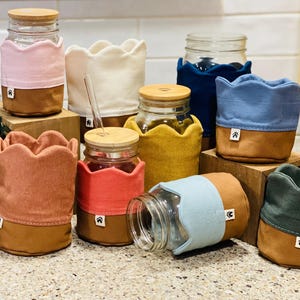 Op de afbeelding: Een verzameling glazen potten met geschulpte stoffen hoezen in diverse kleuren zoals roze, crème, mosterdgeel, koraal, lichtblauw, marineblauw en donkergroen, elk voorzien van een bamboe deksel en een klein wit merklabel.