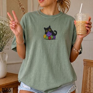Puede incluir: Una camiseta de color verde salvia con un gr&aacute;fico de un gato negro sentado detr&aacute;s de una pila de dados de juego coloridos de varias caras. El gato tiene ojos amarillos grandes y est&aacute; situado en el centro del pecho de la camiseta de cuello redondo.