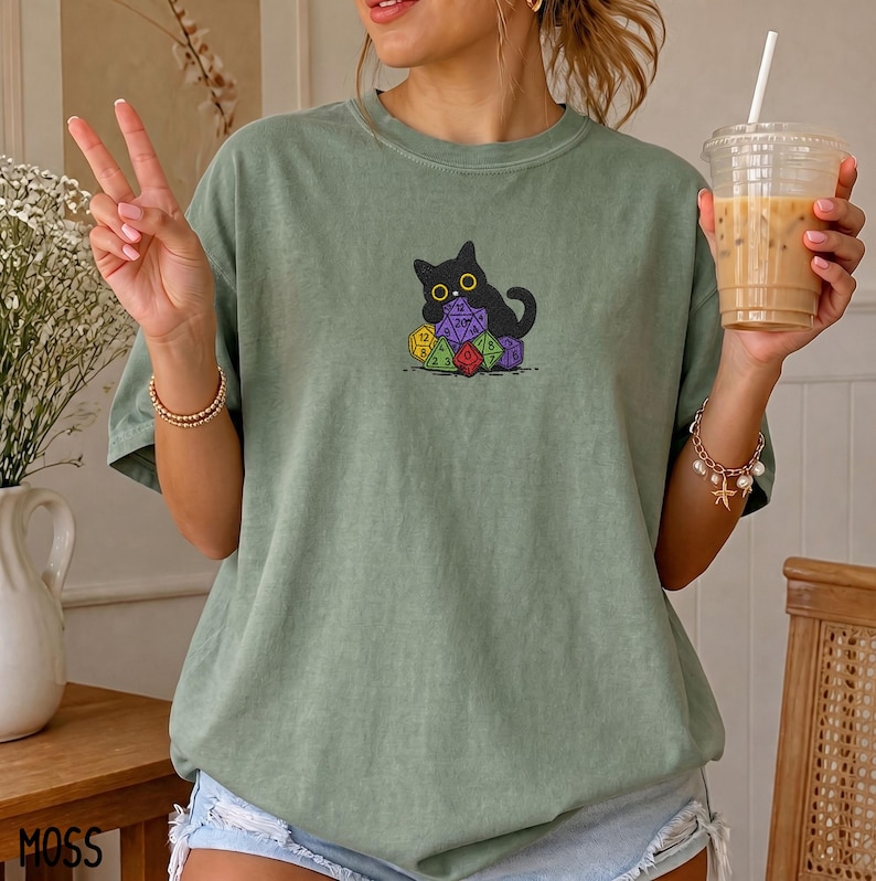 Puede incluir: Una camiseta de color verde salvia con un gr&aacute;fico de un gato negro sentado detr&aacute;s de una pila de dados de juego coloridos de varias caras. El gato tiene ojos amarillos grandes y est&aacute; situado en el centro del pecho de la camiseta de cuello redondo.