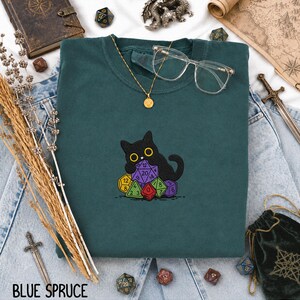 Puede incluir: Una sudadera de cuello redondo en color verde azulado oscuro con un bordado de un gato negro sentado sobre un mont&oacute;n de dados de juego multicolores de varias caras. La prenda incluye un collar con colgante dorado y gafas transparentes.