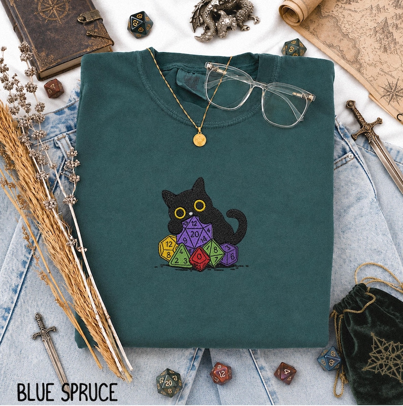 Puede incluir: Una sudadera de cuello redondo en color verde azulado oscuro con un bordado de un gato negro sentado sobre un mont&oacute;n de dados de juego multicolores de varias caras. La prenda incluye un collar con colgante dorado y gafas transparentes.