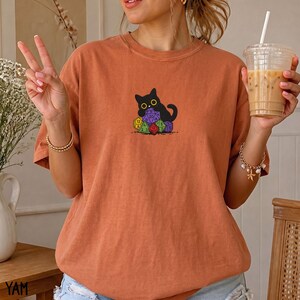 Puede incluir: Una camiseta de manga corta color naranja quemado con un estampado gr&aacute;fico de un gato negro sentado detr&aacute;s de una pila de dados de juego coloridos de varias caras. El gato tiene ojos grandes y est&aacute; centrado en el pecho de la prenda.