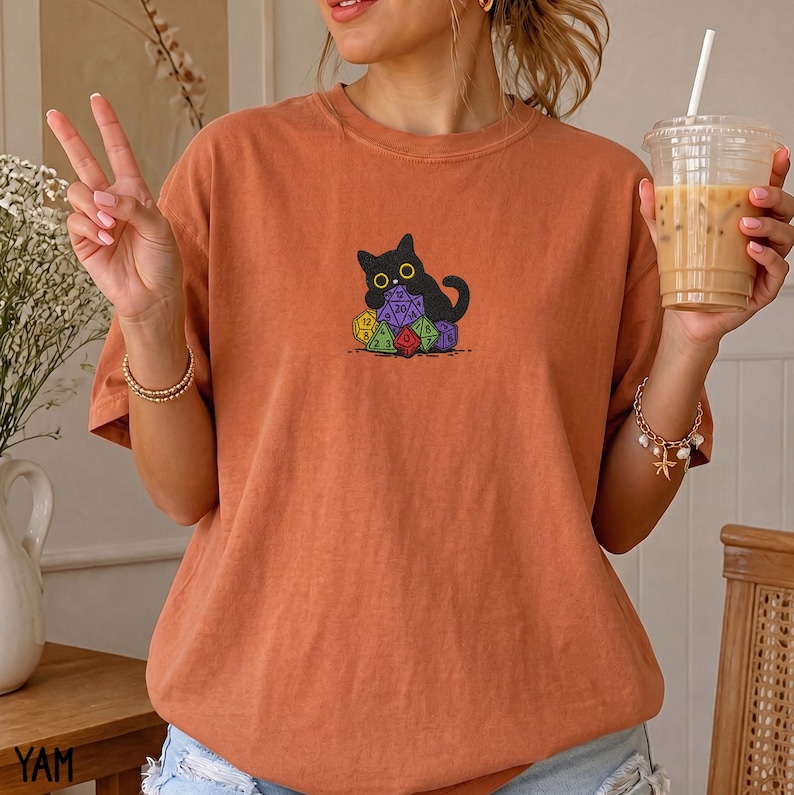 Puede incluir: Una camiseta de manga corta color naranja quemado con un estampado gr&aacute;fico de un gato negro sentado detr&aacute;s de una pila de dados de juego coloridos de varias caras. El gato tiene ojos grandes y est&aacute; centrado en el pecho de la prenda.