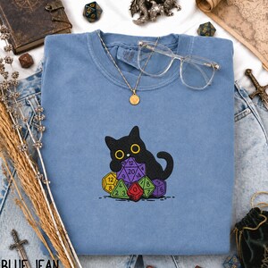 Puede incluir: Una sudadera azul de cuello redondo con un dise&ntilde;o bordado de un gato negro sentado detr&aacute;s de un mont&oacute;n de dados poli&eacute;dricos de colores. La sudadera est&aacute; decorada con un collar con colgante dorado y gafas de montura transparente sobre la tela.