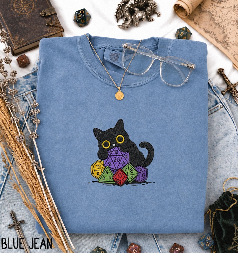 Puede incluir: Una sudadera azul de cuello redondo con un dise&ntilde;o bordado de un gato negro sentado detr&aacute;s de un mont&oacute;n de dados poli&eacute;dricos de colores. La sudadera est&aacute; decorada con un collar con colgante dorado y gafas de montura transparente sobre la tela.