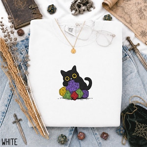 Puede incluir: Una camiseta blanca con un gr&aacute;fico bordado de un gato negro sentado detr&aacute;s de un mont&oacute;n de dados coloridos de varias caras. El gato tiene grandes ojos amarillos. La camiseta tiene una etiqueta Comfort Colors en el cuello interior.