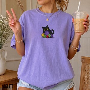 Puede incluir: Una camiseta de manga corta de color morado claro con un gr&aacute;fico de un gato negro sentado detr&aacute;s de una pila de dados de juego de varias caras de colores. El gato tiene ojos amarillos grandes y est&aacute; situado en el centro del pecho.