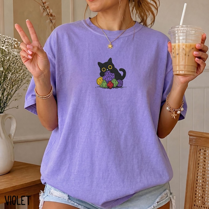 Puede incluir: Una camiseta de manga corta de color morado claro con un gr&aacute;fico de un gato negro sentado detr&aacute;s de una pila de dados de juego de varias caras de colores. El gato tiene ojos amarillos grandes y est&aacute; situado en el centro del pecho.