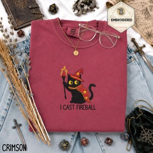 May include: A crimson crewneck sweatshirt featuring an embroidered black cat wearing a red wizard hat and cape with yellow stars, holding a flaming torch. Below the cat, the text reads I CAST FIREBALL in black, sans-serif capital letters.