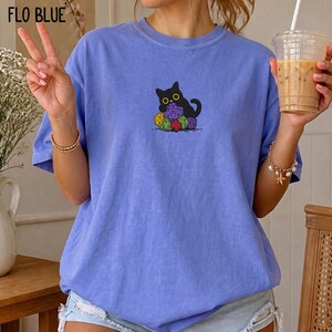 Puede incluir: Una camiseta de color azul b&iacute;garo con un gr&aacute;fico central de un gato negro sentado detr&aacute;s de un mont&oacute;n de dados poli&eacute;dricos de colores. El gato tiene grandes ojos amarillos y est&aacute; situado directamente sobre el grupo de dados.