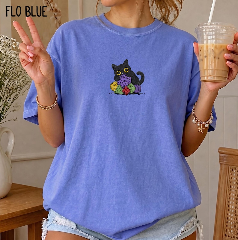Puede incluir: Una camiseta de color azul b&iacute;garo con un gr&aacute;fico central de un gato negro sentado detr&aacute;s de un mont&oacute;n de dados poli&eacute;dricos de colores. El gato tiene grandes ojos amarillos y est&aacute; situado directamente sobre el grupo de dados.