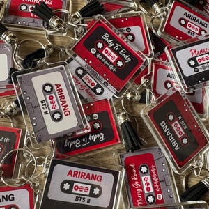 May include: A collection of rectangular acrylic keychains shaped like cassette tapes. Each keychain features a black tassel and displays various text including Arirang, BTS M, Body To Body, Please, and Merry Go Round, along with graphic sound waves.