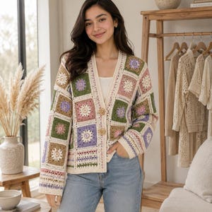 May include: A long-sleeve cardigan constructed from multi-colored crochet granny squares in shades of sage green, lavender, dusty pink, and cream. The garment features a V-neckline, two decorative buttons at the center front, and ribbed cuffs.