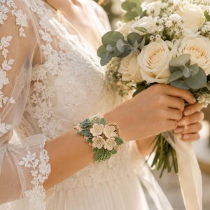 Puede incluir: Una pulsera de ganchillo con flores tridimensionales de color crema y hojas de color verde salvia. El accesorio está elaborado con un intrincado trabajo de aguja, mostrando un patrón floral que envuelve la muñeca con una apariencia texturizada.