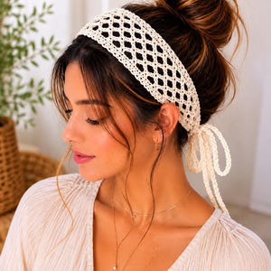 May include: A cream-colored crochet headband featuring an open diamond-shaped lattice pattern. The headband is constructed from yarn and includes two long, thin crocheted ties at the back that hang down, allowing for an adjustable fit around the head.