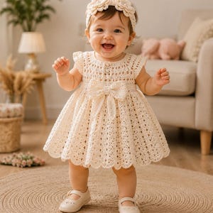 May include: A cream-colored crochet dress with a scalloped hem, a central bow at the waist, and a matching crochet headband featuring two floral accents. The dress has short ruffled sleeves and a button-up bodice, paired with cream-colored shoes.