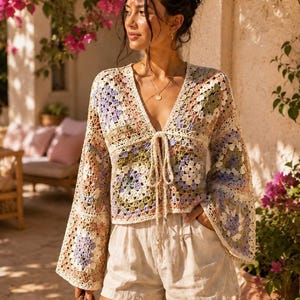 May include: A long-sleeved crochet top featuring a multi-colored granny square pattern in shades of lavender, sage green, and cream. The garment has a deep V-neckline with a central tie closure and wide, flared sleeves, paired with beige linen shorts.