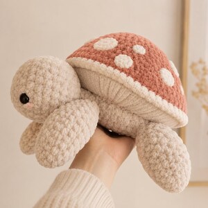 May include: A plush crochet turtle toy featuring a soft beige head and limbs with a textured, mushroom-inspired shell. The shell is a dusty rose colour with white circular spots and a cream-coloured underside. It has small black plastic eyes.