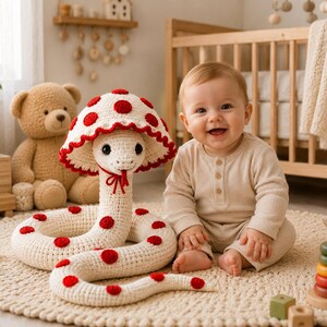 May include: A crocheted snake toy featuring a cream-colored body with red circular spots and a matching mushroom-shaped hood. The snake has a small red tie under its chin, a red tongue, and dark eyes, crafted with a textured yarn finish.