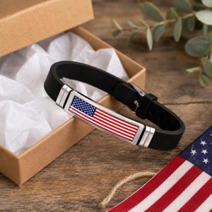 May include: A black silicone bracelet featuring a rectangular metal plate with a printed American flag design. The bracelet has a silver-toned metal buckle and is positioned inside an open cardboard gift box lined with white tissue paper.