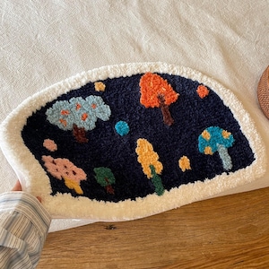 Puede incluir: Una alfombra de mechones semicircular con un fondo azul marino oscuro y un borde blanco. La alfombra muestra un diseño de paisaje colorido con varios árboles y setas en tonos naranja, azul claro, amarillo, rosa y verde.
