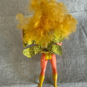 May include: A doll with voluminous curly yellow hair, wearing a pair of large, glittery gold wings attached to the back. The doll has peach-colored legs and wears bright yellow boots, with a yellow piece visible at the waist area.