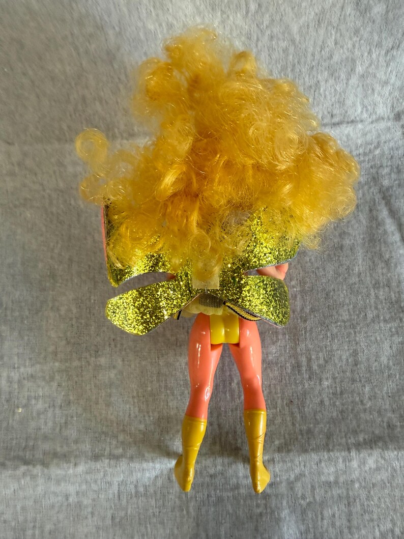 May include: A doll with voluminous curly yellow hair, wearing a pair of large, glittery gold wings attached to the back. The doll has peach-colored legs and wears bright yellow boots, with a yellow piece visible at the waist area.