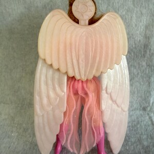 May include: A doll figure viewed from the back, featuring large, pale, pearlescent plastic wings attached to a circular emblem at the neck. The doll wears a sheer, light pink fabric skirt and bright pink boots, with brown hair visible above the wings.